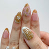 Hand with decorated nails featuring floral and geometric patterns, wearing a gold ring and pearl earrings on a white background.